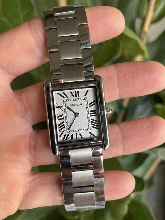Silver watch with a rectangular face held in a hand against a green leafy background. Sanda 1108.